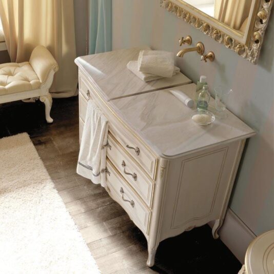 Designer Italian Bathroom Cabinet with Sliding Top Elegant bathroom with a vintage vanity featuring a marble countertop, rolled towels, a soap dispenser, and a toothbrush. The Designer Italian Bathroom Cabinet with Sliding Top adds sophistication to the space. The vanity has ornate drawers with metallic handles, and a large framed mirror hangs above. A plush, cushioned seat and a fluffy rug are also visible.