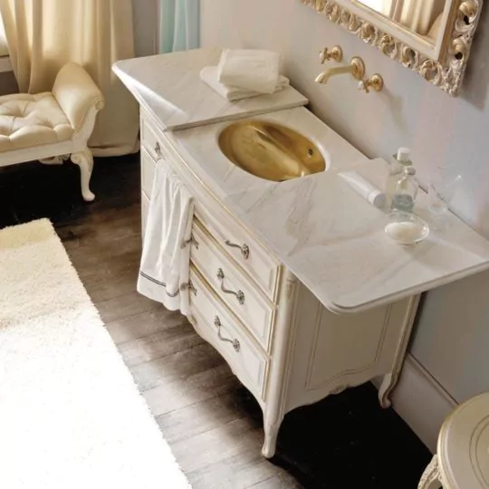 A luxurious bathroom featuring a Designer Italian Bathroom Cabinet with a sliding top and an ornate vanity with a gold sink set in a marble countertop. The vanity has detailed drawers and a plush towel hanging from it. Above, a decorative mirror adorns the wall. Additionally, the room boasts a cushioned bench and a soft white rug for added comfort.