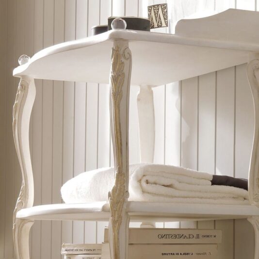 A close-up of the Designer Italian Freestanding Bathroom Shelves, a vintage-styled white wooden side table with ornate legs and two shelves. Perfect for any bathroom, the upper shelf showcases small decorative items, while the lower shelf neatly accommodates a folded white towel and books. The background features vertical white paneling.