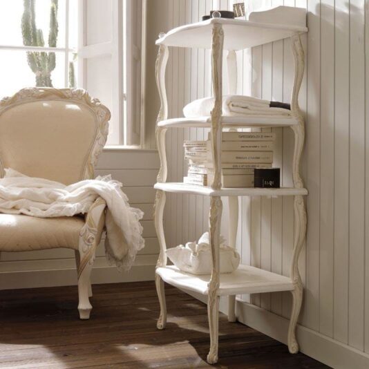 The Designer Italian Freestanding Bathroom Shelves stand against a wooden-paneled wall in a cozy room. These vintage-style shelving units hold folded towels, a stack of books, and a teapot. To the left, an ornate cream armchair is draped with a blanket. A nearby window lets in natural light.
