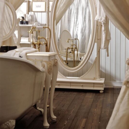 A vintage-style bathroom featuring a free-standing white bathtub with claw feet, a large oval standing mirror, and an elegant vanity table with ornate legs. Brass fixtures adorn the space, complemented by a Designer Italian Rococo Console Table with Mirror. White wooden paneling covers the walls, and the floor is dark hardwood.