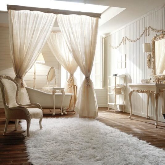 A light and airy vintage-style room features a white canopy bed, an elegant armchair, and an ornate vanity. Light curtains surround the bed, and a fluffy white rug covers the wooden floor. The walls are adorned with white paneling, complemented by a Designer Italian Rococo Console Table with mirror above it.