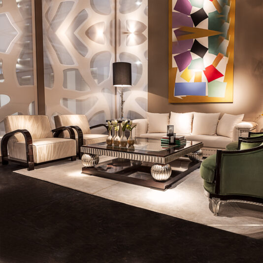 Ebony And Silver Square Coffee Table A modern living room features a beige sofa, two cream-colored armchairs with dark wooden arms, and an olive green armchair. The Ebony And Silver Square Coffee Table sits on a white rug. A floor lamp and abstract wall art with colorful geometric shapes complete the space.
