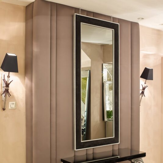 Elegant Contemporary Nubuck Black Mirror A modern, elegant interior features an Elegant Contemporary Nubuck Black Mirror with a dark frame mounted on a padded wall panel. Two black wall sconces with white shades are symmetrically placed on either side of the nubuck-backed mirror, adding a touch of refinement to the beige-colored room.