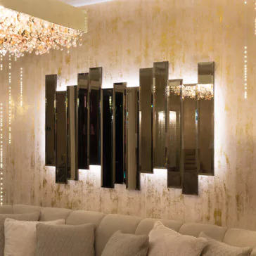 Exclusive Backlit Bronze Mirror A contemporary living room highlights the Exclusive Backlit Bronze Mirror, distinguished by its large, staggered vertical panels and set against a textured golden wall. Overhead, an elegant chandelier with dangling, sparkling crystals enhances the space. A plush sofa below the mirror is adorned with light gray cushions.