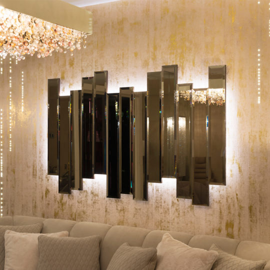A contemporary living room highlights the Exclusive Backlit Bronze Mirror, distinguished by its large, staggered vertical panels and set against a textured golden wall. Overhead, an elegant chandelier with dangling, sparkling crystals enhances the space. A plush sofa below the mirror is adorned with light gray cushions.