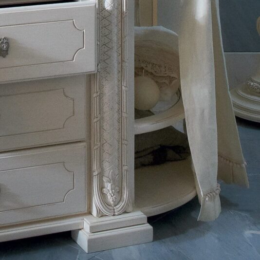 Exclusive Italian 4 Drawer Bathroom Cabinet A close-up of the Exclusive Italian 4 Drawer Bathroom Cabinet in a soft cream color. This vintage-style dresser showcases ornate carvings and a partially hidden tiered shelf holding a ball-shaped object and folded fabric. To the right, a beige curtain drapes down, partially visible.