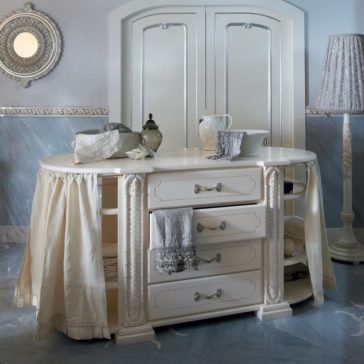 A vintage-style vanity with a white, ornate design and a large mirror featuring draped fabric on the sides. It showcases decorative handles and displays various items like a teapot, folded linens, and trays positioned both on top and within its compartments. Nearby stands an Exclusive Italian 4 Drawer Bathroom Cabinet accompanied by a tall lamp and an intricate wall mirror.