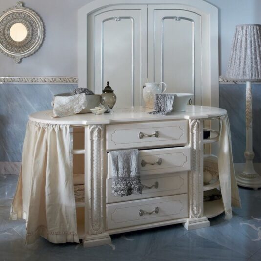 A vintage-style vanity with a white, ornate design and a large mirror featuring draped fabric on the sides. It showcases decorative handles and displays various items like a teapot, folded linens, and trays positioned both on top and within its compartments. Nearby stands an Exclusive Italian 4 Drawer Bathroom Cabinet accompanied by a tall lamp and an intricate wall mirror.