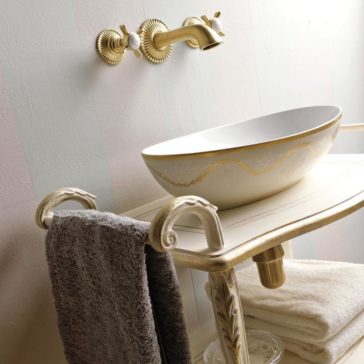 A luxurious bathroom setup features the Exclusive Italian Bathroom Basin Console with a white and gold vessel sink atop an ornate vanity table adorned with carved details. Above the sink, a decorative gold faucet with wall-mounted handles adds elegance. A soft gray towel hangs on the side of the console, while white towels are neatly stacked below.
