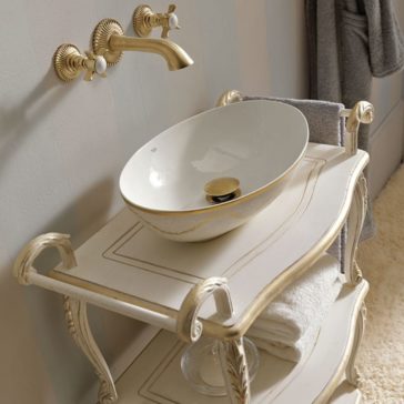 The Exclusive Italian Bathroom Basin Console features a vintage-style white and gold ceramic vessel basin on an ornate, cream-colored vanity stand. This elegant piece boasts two lower shelves for holding folded towels and is complemented by a curved golden faucet with matching handles mounted on the wall above the basin.
