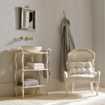 An elegant bathroom with vintage decor features the Exclusive Italian Bathroom Basin Console. An ornate mirror hangs above the small sink, which sits atop the decorative stand with shelves holding towels. A grey robe is on a wall hook, and a cushioned, vintage-style chair rests nearby on a light carpet. The walls are adorned with soft vertical stripes.