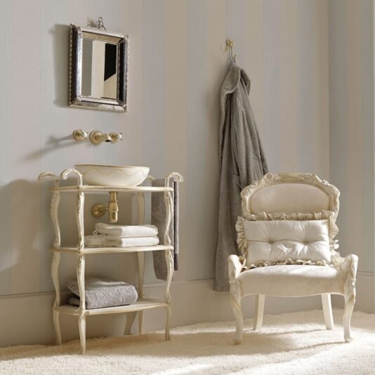 An elegant bathroom with vintage decor features the Exclusive Italian Bathroom Basin Console. An ornate mirror hangs above the small sink, which sits atop the decorative stand with shelves holding towels. A grey robe is on a wall hook, and a cushioned, vintage-style chair rests nearby on a light carpet. The walls are adorned with soft vertical stripes.