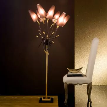 Exclusive Italian Bronze Lacquered Floor Lamp An Exclusive Italian Bronze Lacquered Floor Lamp with multiple angled lampshades emits a warm light, positioned next to a high-backed white chair. A stack of books and a gold bowl are placed on the seat of the chair. The background features a dark wall and a textured backdrop.