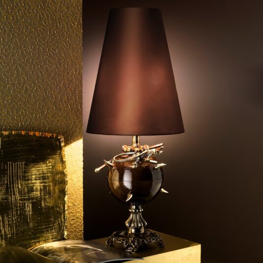 The Exclusive Italian Bronze Table Lamp boasts a contemporary design with a sleek black lampshade and an elegant, ornate base. The base is highlighted by a dark glossy sphere embellished with metallic flourishes and intricate details. Adding to its appeal, this exceptional lamp is showcased on a gold-colored table against a textured, patterned wall.
