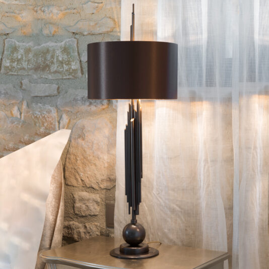 An Exclusive Italian Brown And Gold Table Lamp with a black cylindrical shade and a unique vertical metal rod design sits on a metallic round base. The lamp rests on a small table next to a stone wall and sheer white curtain, with part of a sofa visible in the background.