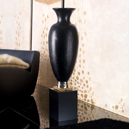 The Exclusive Italian Designer Ornamental Black Vase, with a textured finish, stands on a square pedestal in an elegant modern room accented by a fur throw on a dark sofa and a patterned cream and gold wall.