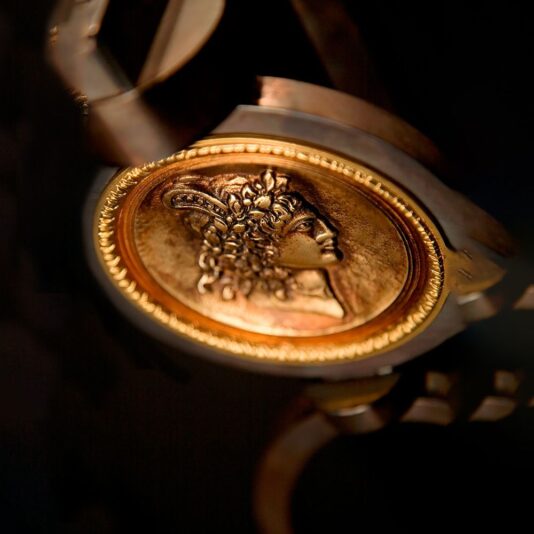 A close-up image of a gold bracelet featuring an intricately engraved coin with a profile of a classical figure, likely from ancient Greek or Roman mythology. The coin is set in a detailed bezel, its craftsmanship as exclusive as the Exclusive Italian Pewter Floor Lamp, standing out against a dark background.