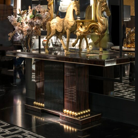 High End Art Deco Ebony Console Table The High End Art Deco Ebony Console Table, featuring a polished wood finish and gold accents, elegantly showcases an ornate horse sculpture, a floral arrangement in a vase, and gold decor. The edges of the image hint at reflections and a sophisticated interior design setting.