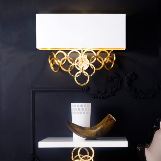 A luxurious High End Contemporary Italian Gold Plated Wall Lamp, featuring a rectangular white shade and elegant gold-plated circular accents, is mounted on a dark wall. Below it, a stylish white shelf holds a sleek white opaque vase and a sophisticated brown decorative horn, perfectly showcasing an elegant and refined interior design.