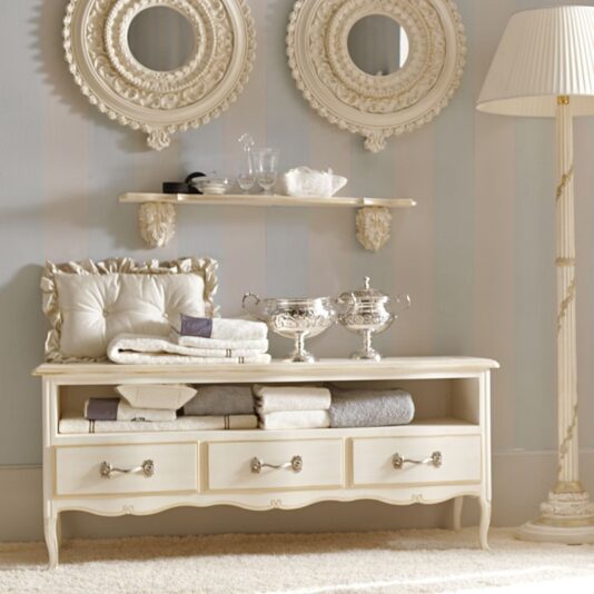 A vintage-style vanity area showcases an ivory-colored wooden dresser with four drawers. Above it, the Luxury Ivory and Gold Italian Wall Fixing Shelf elegantly displays glassware between two ornate round mirrors. Cushions and folded towels rest on the dresser, accented with silverware. To the right, a tall decorative floor lamp adds height and charm to the space.