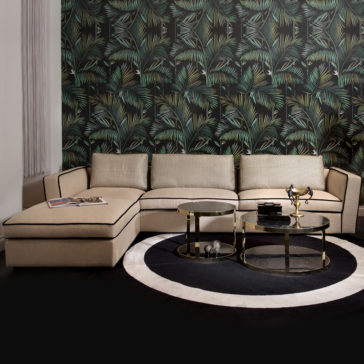 Italian Designer Modern Modular Corner Sofa A modern living room showcasing an Italian Designer Modern Modular Corner Sofa set against tropical leaf-patterned wallpaper. The space features two round coffee tables with gold frames and glass tops, positioned on a black and white circular rug. Magazines and decorative items are stylishly arranged on the tables and sofa.