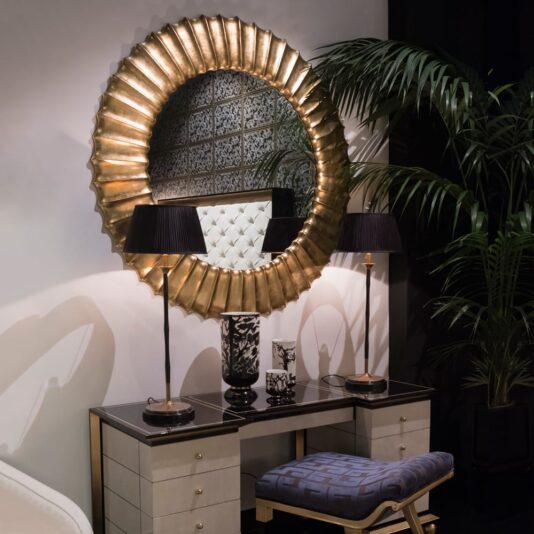Elegant vanity setup featuring the exquisite Large Round Gold Designer Mirror above a stylish black and white desk with drawers. Two sophisticated black-shaded lamps flank a decorative black and white vase on the desk. Beneath, a patterned stool adds character while green plants flourish nearby.