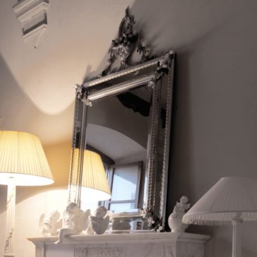 A Luxurious Venetian Italian Mirror With Semi Precious Stones hangs above a white marble mantel, with elegant pleated-shade lamps and ceramic figurines arranged below, creating a cozy, classic look.