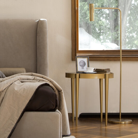 A cozy bedroom showcases a Luxury Designer Italian Leather Bed With Walnut Veneer Headboard, elegantly draped in beige bedding. A small gold side table holds a framed photo and a white teacup on a saucer. Nearby, a gold floor lamp illuminates the space, while a large window with a wooden frame reveals lush greenery outside.
