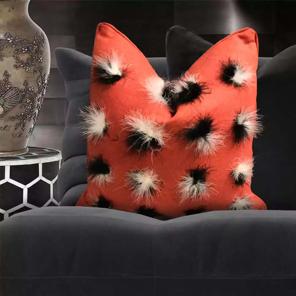 A plush orange pillow with a pattern of black and white feathers rests on a dark gray velvet sofa, adding a touch of cosy luxury to the setting. A decorative vase with floral designs sits elegantly on a table in the background.