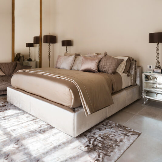 A modern bedroom showcasing a Luxury Italian Ivory Velvet Upholstered Bed, adorned with multiple pillows and a neatly arranged duvet. The room includes mirrored nightstands with matching brown lamps on either side, and a patterned area rug partially covering the tiled floor.