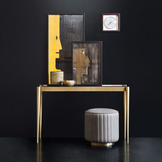 A Luxury Italian Marble Brass Designer Console Table with a gold finish displays two framed abstract art pieces, gold bowls, and a candle holder. Below is a round grey pouffe, while a small wall clock hangs on the black wall behind.