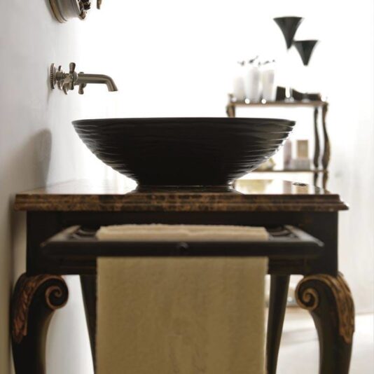 Marble Topped Designer Italian Bathroom Basin A chic bathroom features a *Marble Topped Designer Italian Bathroom Basin* positioned gracefully atop an ornate wooden vanity. This vanity, adorned with intricate carvings, also includes a lower shelf that holds a folded white towel. In the background, a matching shelf elegantly displays various toiletries and decor items. A sleek open faucet is mounted on the wall.
