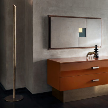 Minimal Contemporary Italian Burnished Brass Floor Lamp A modern interior includes the Minimal Contemporary Italian Burnished Brass Floor Lamp, an orange and gold chest of drawers, a small horse figurine, black stacked shapes, and minimalist geometric artwork on a textured grey wall.