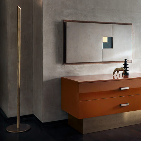 A modern interior includes the Minimal Contemporary Italian Burnished Brass Floor Lamp, an orange and gold chest of drawers, a small horse figurine, black stacked shapes, and minimalist geometric artwork on a textured grey wall.