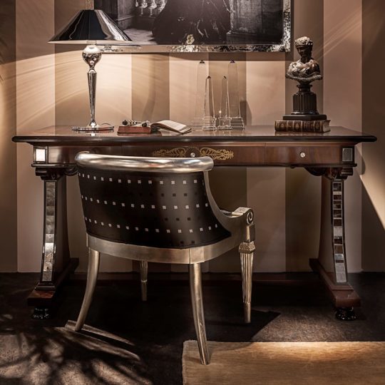 The Neoclassical Mahogany Writing Desk And Chair create a classic workspace, featuring ornate details and paired with books, a notepad, two glass obelisks, and a lamp against elegant striped walls.