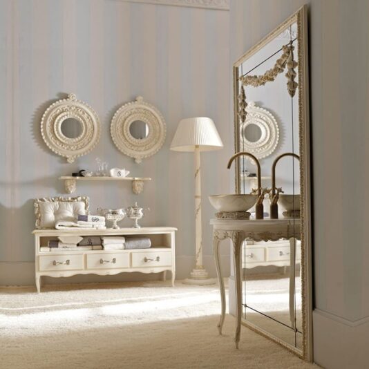 A luxurious bathroom showcases an Opulent Italian Bathroom Console with Mirror, alongside two round decorative mirrors mounted on the wall. It also includes a sleek table featuring twin sinks and two golden faucets, complemented by a white bench adorned with pillows and folded towels. A standing lamp with a pleated shade casts a warm glow, all set against an elegant backdrop of light striped wallpaper.