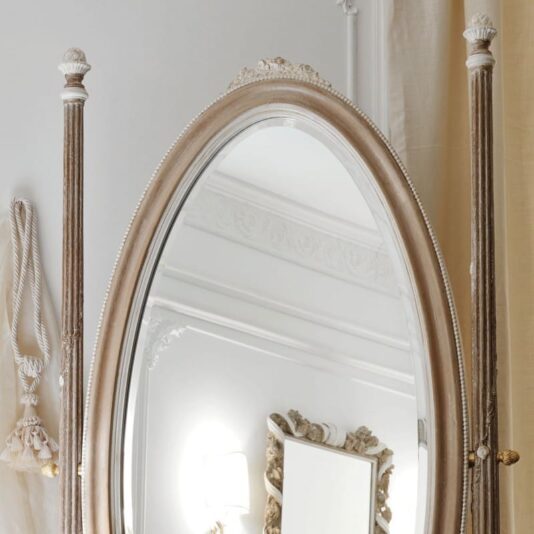 Ornate Wooden Italian Oval Dressing Mirror The Ornate Wooden Italian Oval Dressing Mirror features a beautifully carved frame and sits on a decorative stand. It reflects part of a white room adorned with elegant molding and a wall-mounted light fixture, while a tasseled rope hangs on the wall to the left of the mirror.