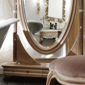 Ornate Wooden Italian Oval Dressing Mirror An elegant bathroom features the Ornate Wooden Italian Oval Dressing Mirror with a decorative frame. Reflections show a freestanding bathtub, a vintage-style vanity with a matching mirror, and white walls adorned with intricate moldings. A cushioned chair with a wooden frame is in the foreground.