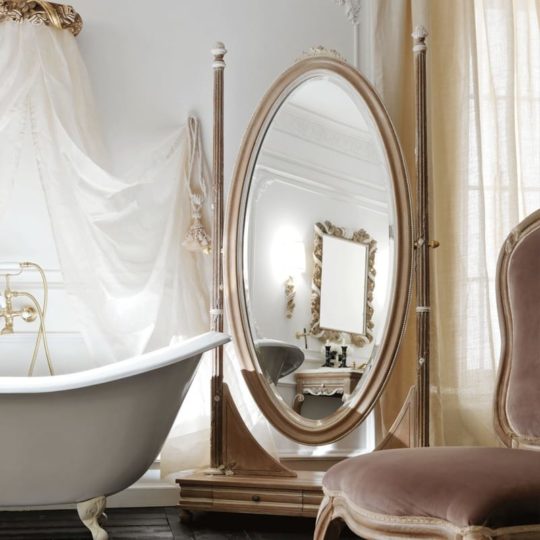 A luxurious bathroom with a freestanding bathtub and the Ornate Wooden Italian Oval Dressing Mirror. The room features elegant drapery, a plush chair with fabric upholstery, and intricate wall decor, all in a sophisticated, light color palette.