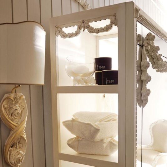 Reproduction Italian Freestanding Bathroom Mirror A cozy spa-like interior featuring a white shelf adorned with ornate carvings. The shelf holds stacked white towels, a glass bowl with white rounds, and dark-colored containers. A decorative wall sconce and the Reproduction Italian Freestanding Bathroom Mirror accentuate the elegant ambiance.
