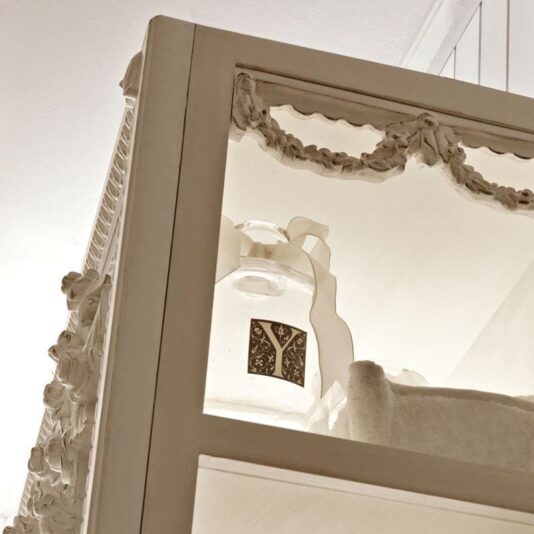 Reproduction Italian Freestanding Bathroom Mirror Close-up view of a decorative white cabinet with intricate carvings and detailing. Inside, there's a glass bottle with a floral design on its label. The vintage and elegant style of the cabinet complements the Reproduction Italian Freestanding Bathroom Mirror placed beside it.