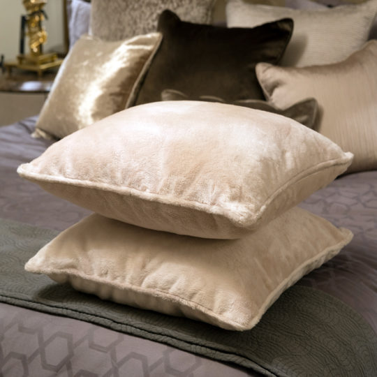 A Set Of 2 Italian Soft Touch Designer Scatter Cushions in beige is stacked on a neatly made bed with neutral pillows and a textured grey blanket, while gold décor and more cushions enhance the stylish setting.
