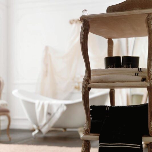 A luxurious bathroom featuring Wooden Italian Freestanding Bathroom Shelves adorned with stacked towels and two black candles, alongside a white clawfoot bathtub and sheer curtains, exudes elegance and serenity with a hint of classic design.