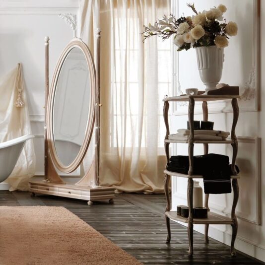 A sophisticated bathroom features an ornate oval standing mirror, a white bathtub, elegant sheer curtains, and Wooden Italian Freestanding Bathroom Shelves holding black and white towels. A tall vase with white flowers tops the shelves, enhancing the ambient, classic decor.