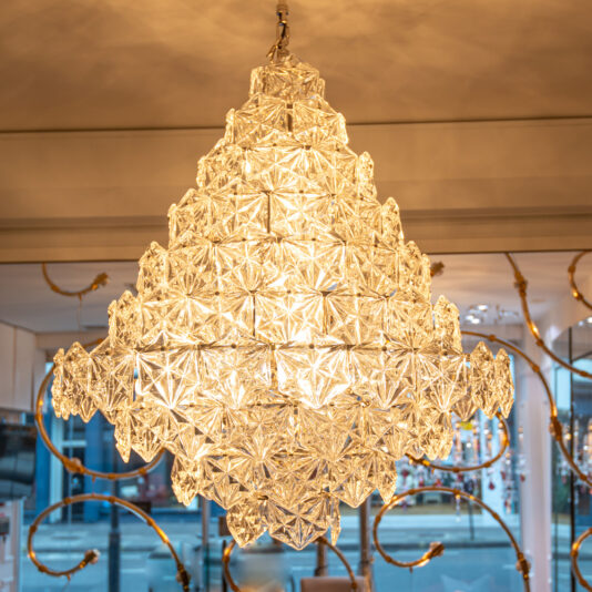 The Large Faceted Glass Pendant Chandelier glows warmly from the ceiling in an elegant room with golden accents and large windows, highlighting its intricate geometric glass design.