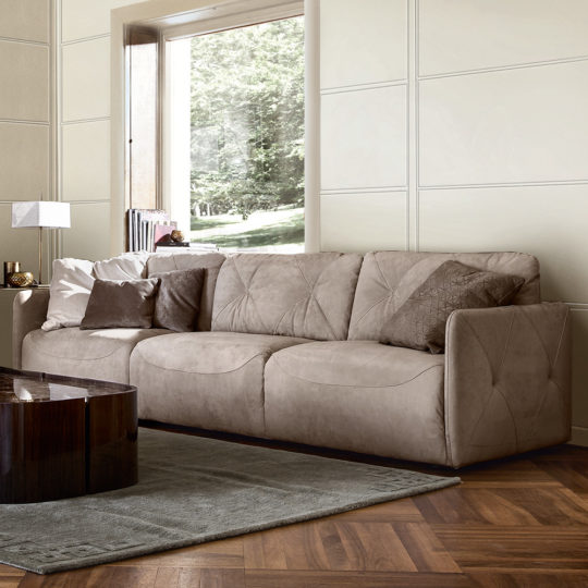 The Luxury Nubuck Leather Designer Italian Sofa with patterned cushions is placed on a grey rug in a modern living room featuring wooden floors, neutral walls, a large window, and a dark wood coffee table in front.