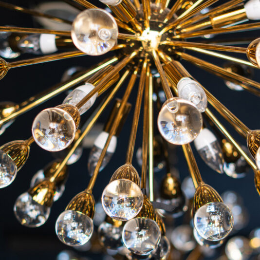 Close-up of the Striking Round Contemporary Chandelier showcasing its starburst design. Gold rods extend outward, each ending in a clear, round bulb against a dark background.