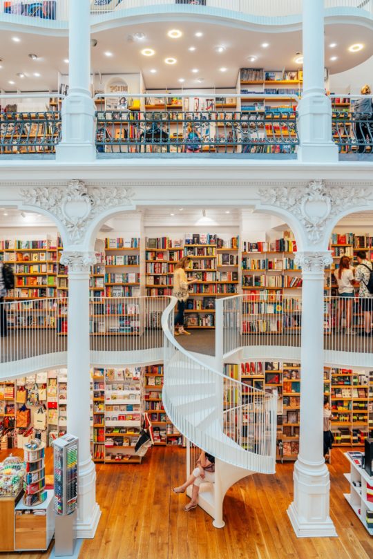 A bright, multi-level bookstore with white columns and decorative arches beckons visitors. The main floor and mezzanine levels, each a charming book nook filled with shelves, are connected by a graceful white spiral staircase. Shoppers browse books while warm wooden floors contrast with the pristine decor.