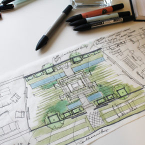 An architectural landscape design blueprint laid out on a desk with various pens, markers, and a notebook nearby. The sketch includes hand-drawn details and notes, showing pathways, greenery, and structural elements of an outdoor retreat, all accompanied by color highlights.